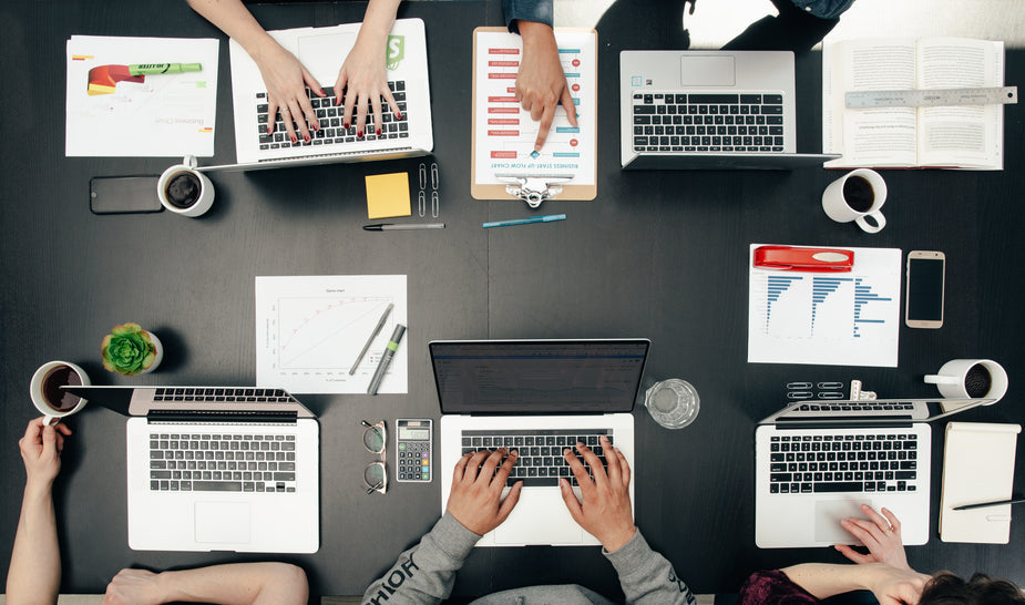 Small group of people working at a table with laptops, printed charts, and coffee.
