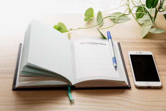 An open notebook on a table with a page titled Top 5 Business Ideas, ready to start planning your new business? A phone lies next to the notebook.